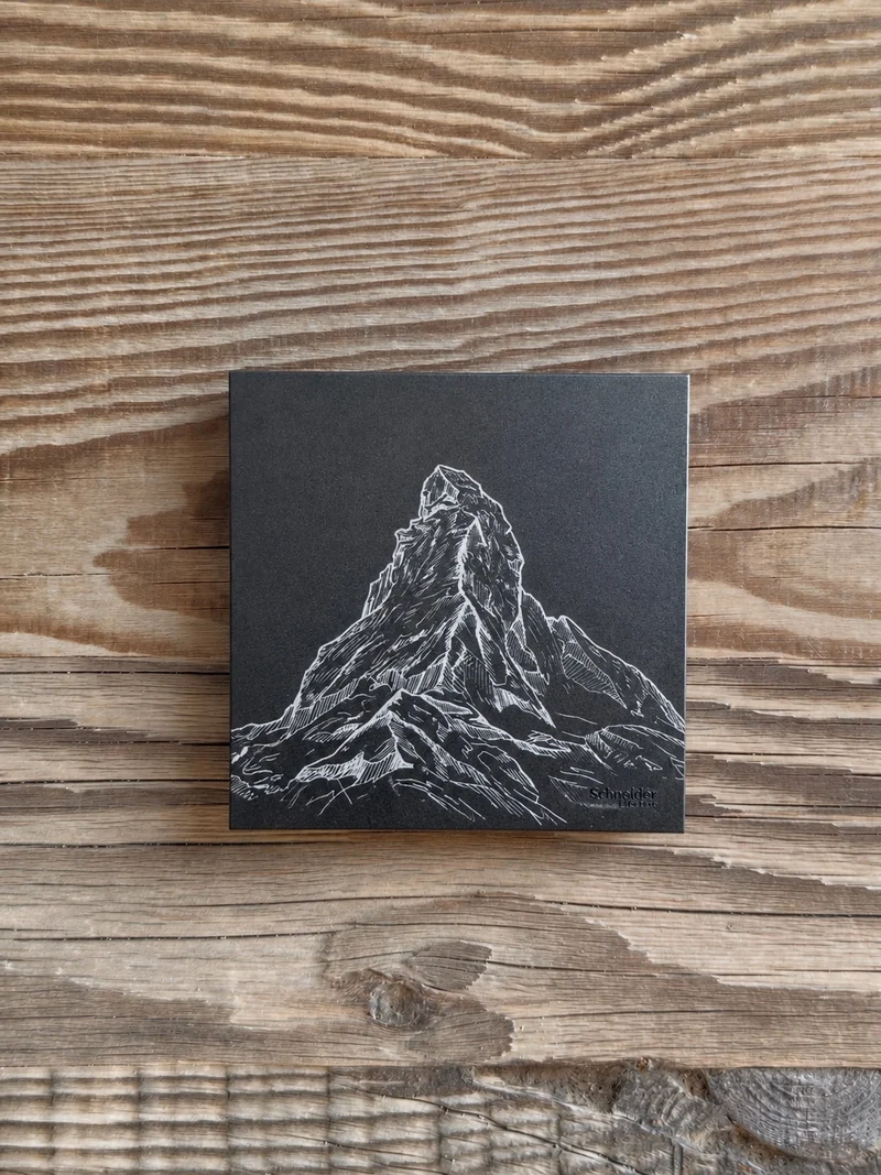 Montagne gravée en traits clairs sur une plaque noire, présentée sur un fond bois.